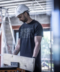 A man wearing a Regatta Professional Pro trade T-shirt and a white hard hat stands indoors at a construction site, holding a wooden plank. A metal ladder and unfinished walls are visible in the background.