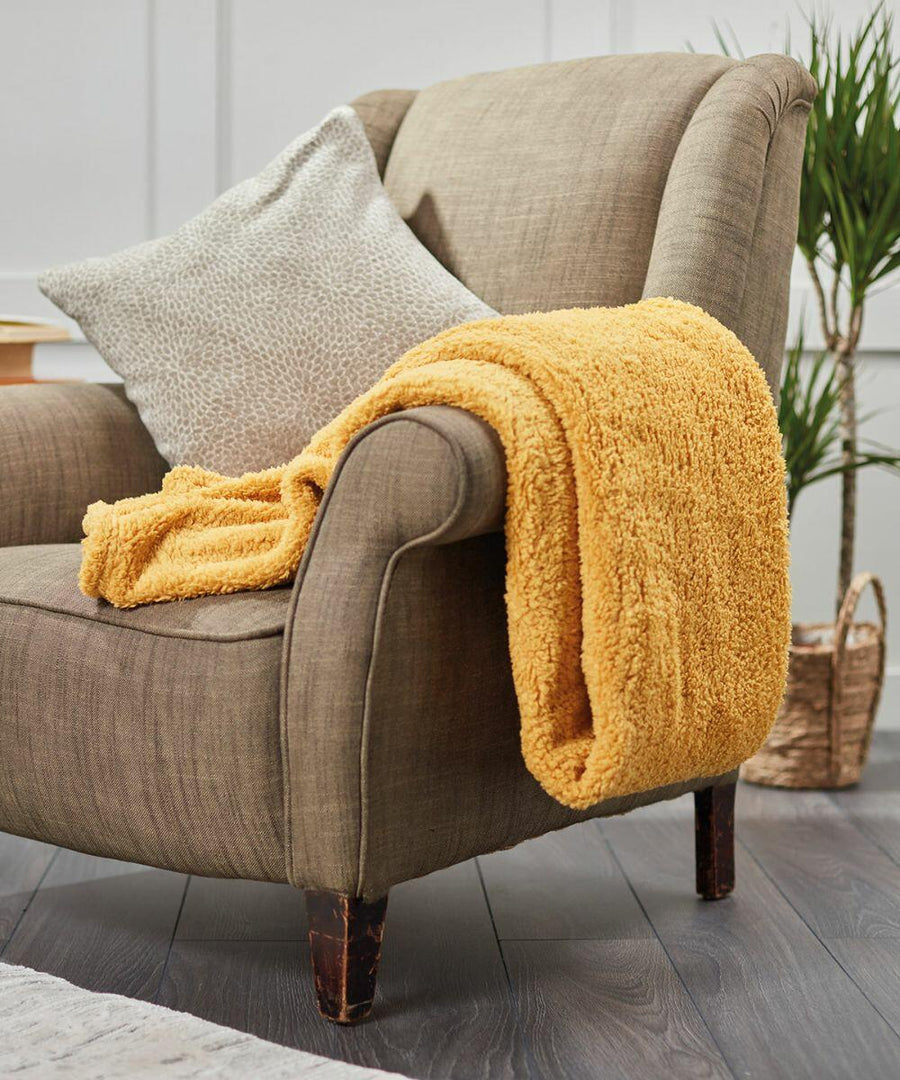 A beige armchair with a pale patterned cushion and a yellow fuzzy blanket draped over the armrest, next to a plant in a woven basket on a gray wood floor, featuring the Ribbon RI010.