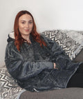 A woman with long red hair sits on a grey sofa, wearing the Ribbon Heated Eskimo blanket hoodie in grey fleece. A patterned grey and white blanket is draped over the sofa, with a plain wall in the background.