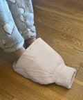 A person in heart-patterned pyjama bottoms has both feet inside a single large Ribbon Luxury foot hot water bottle with a plush faux fur cover, resting on a wooden floor.