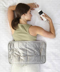 A woman lies on her stomach on a white textured surface, holding a remote while the Ribbon Electric weighted heat pad rests on her lower back.
