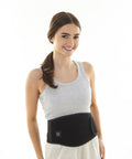 A woman with brown hair in a ponytail, wearing a grey tank top and the Ribbon Heated Waistband, stands against a plain white background and smiles at the camera.