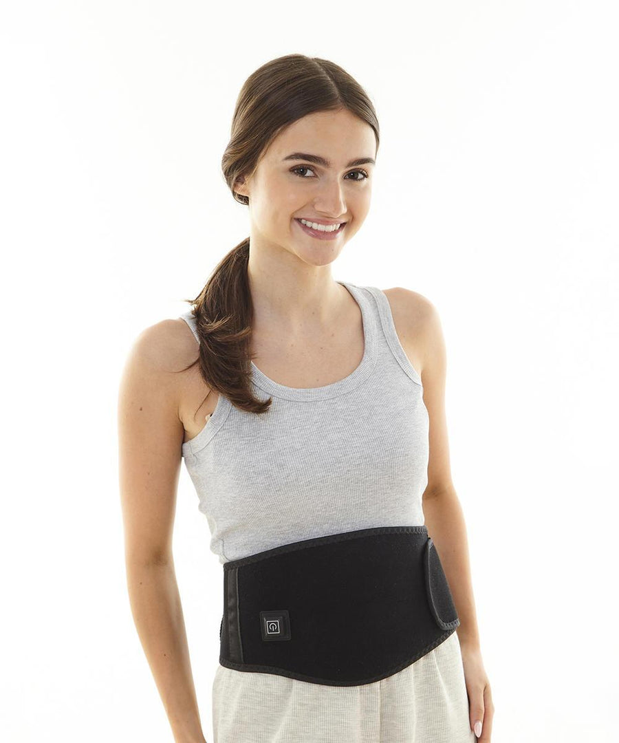 A woman with brown hair in a ponytail, wearing a grey tank top and the Ribbon Heated Waistband, stands against a plain white background and smiles at the camera.