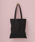 A Nutshell Organic cotton shopper long handle tote in plain black, featuring two handles and a small green side tag, hangs on a wooden peg against a light pink wall.