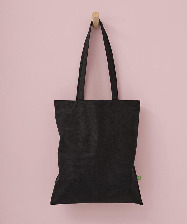 A Nutshell Organic cotton shopper long handle tote in plain black, featuring two handles and a small green side tag, hangs on a wooden peg against a light pink wall.