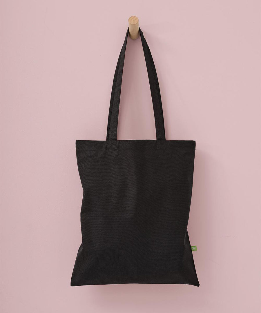 A Nutshell Organic cotton shopper long handle tote in plain black, featuring two handles and a small green side tag, hangs on a wooden peg against a light pink wall.