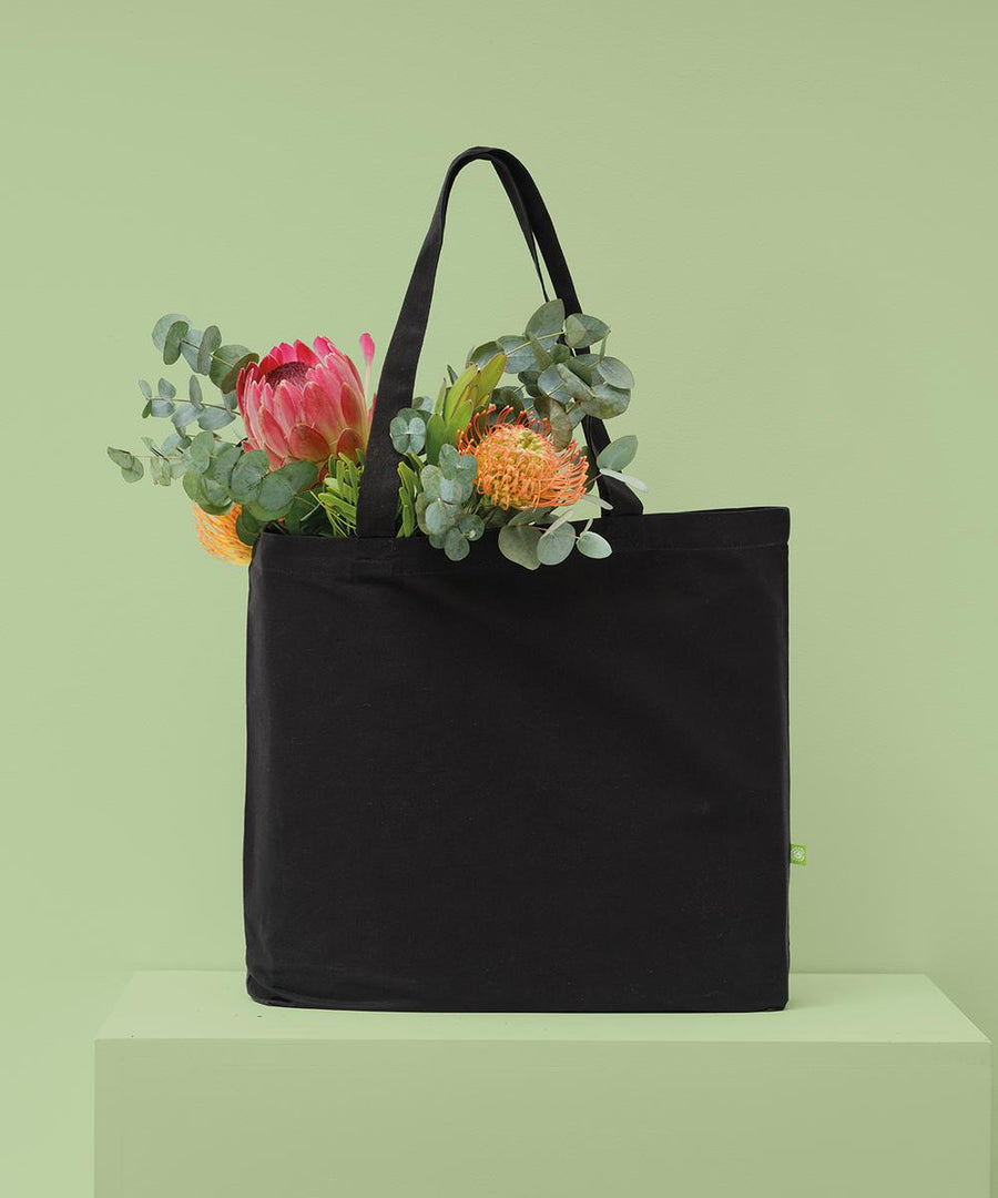 A black organic canvas flat-base landscape shopper by Nutshell with handles sits on a light green surface against a matching green wall, filled with assorted flowers and greenery including eucalyptus and bright blooms.