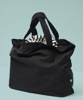 The Nutshell Elba organic canvas premium zip tote in black, featuring short handles, rests on a light blue surface. It’s partially unzipped to reveal its black and white striped interior lining.