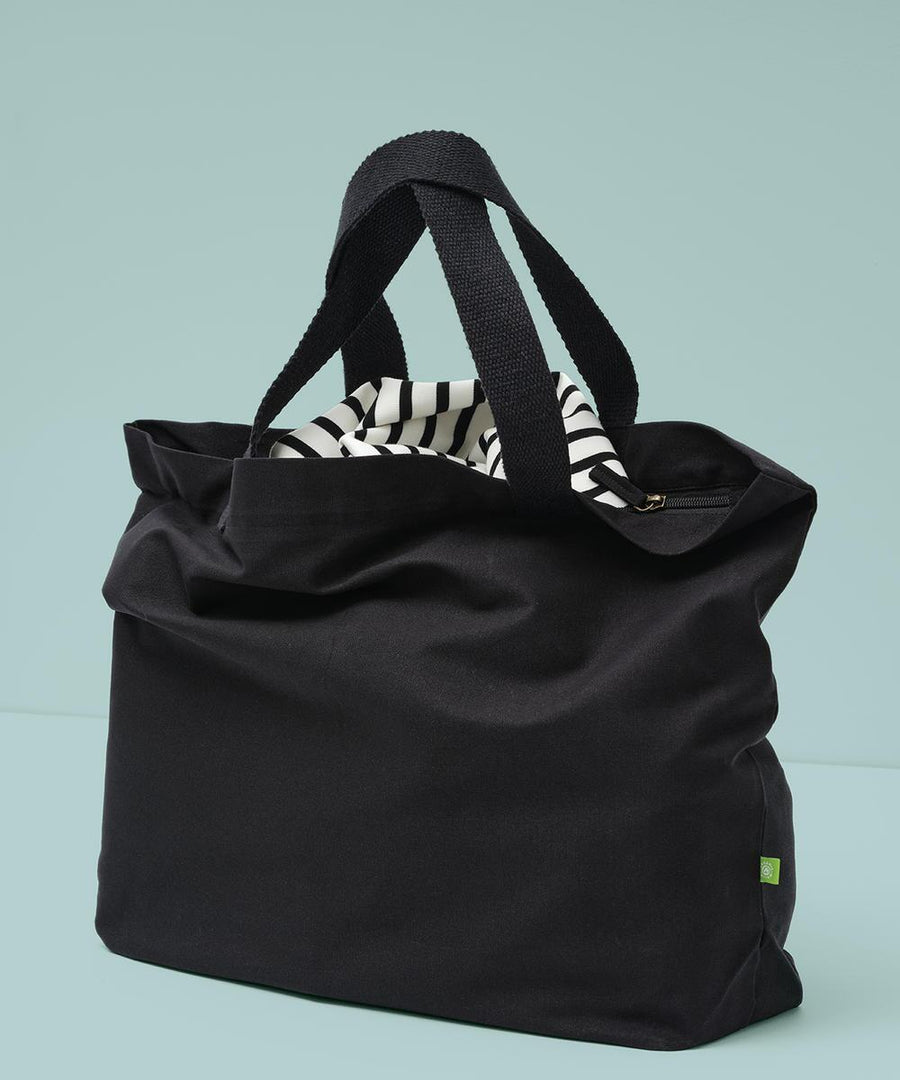 The Nutshell Elba organic canvas premium zip tote in black, featuring short handles, rests on a light blue surface. It’s partially unzipped to reveal its black and white striped interior lining.