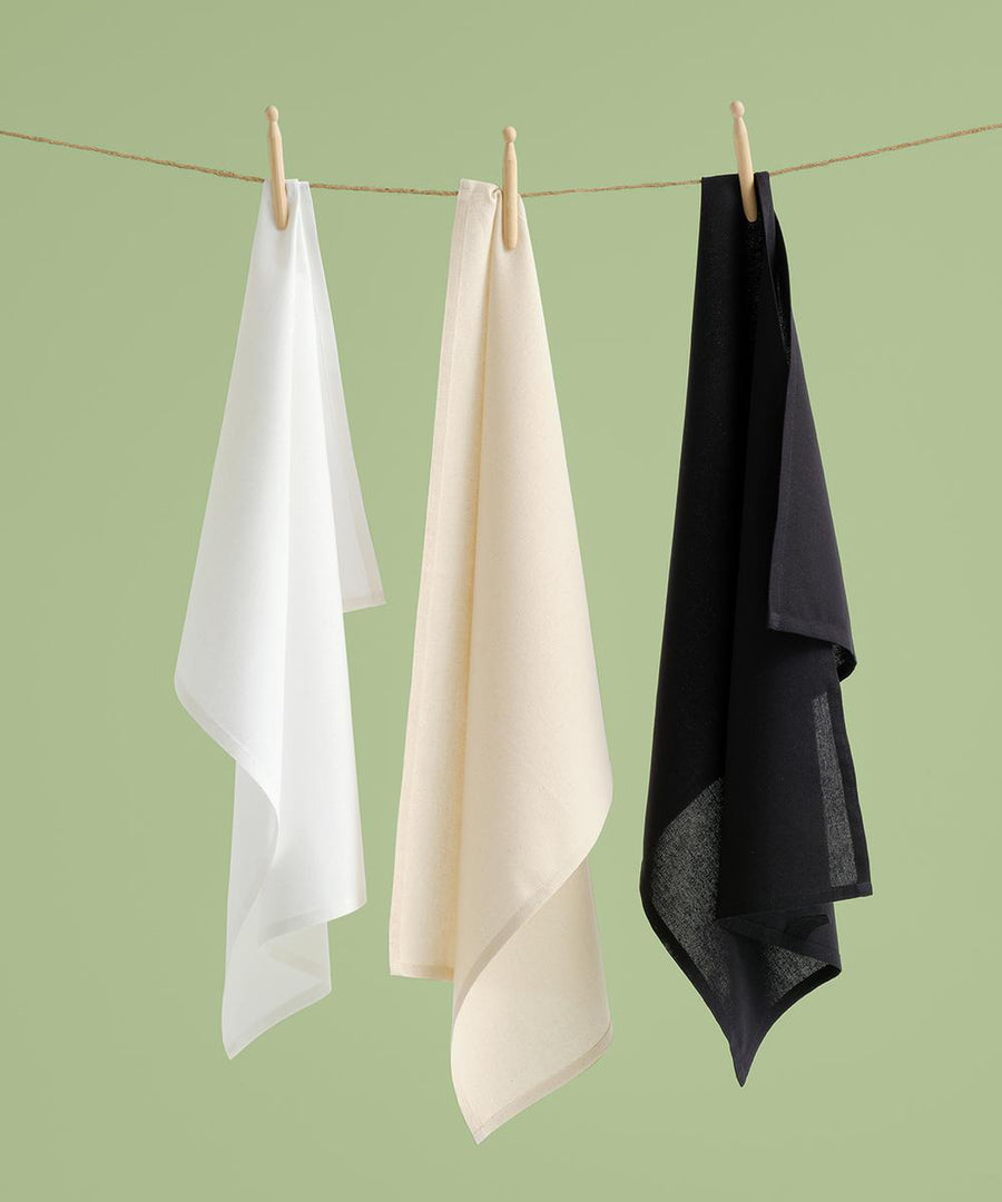 Three organic cotton tea towels by Nutshell—in white, beige, and black—hang side by side on a string with wooden pegs against a light green background.