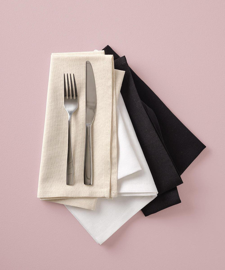 A fork and knife rest on a Nutshell Organic cotton serviette in cream, layered over white and black serviettes, all arranged on a pink surface.