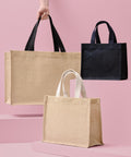 A hand holds a tan Nutshell Jute shopper with black handles. Beside it are a black Jute shopper and another tan one with white handles, all displayed on pink steps against a pink background.