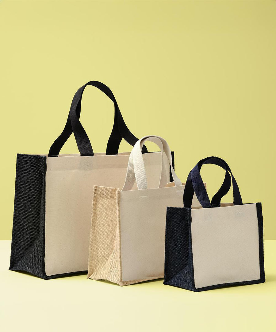 Three Nutshell jute canvas panel shopper bags in various sizes are displayed on a yellow surface with a yellow backdrop. All are beige with black side panels and handles, except the middle one, which features beige handles.