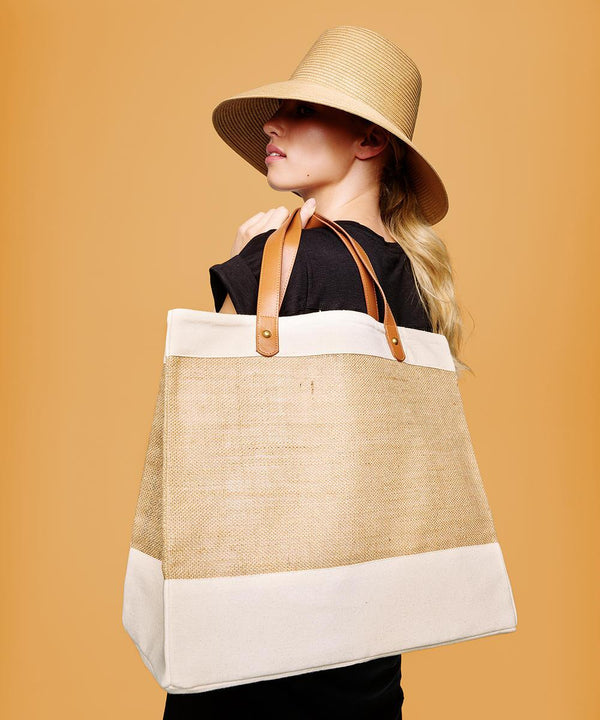 A person in a black shirt and wide-brimmed beige hat carries the Nutshell Adley jute with faux leather tote, featuring tan handles, beige sides, and white trim, standing against a solid light brown background.