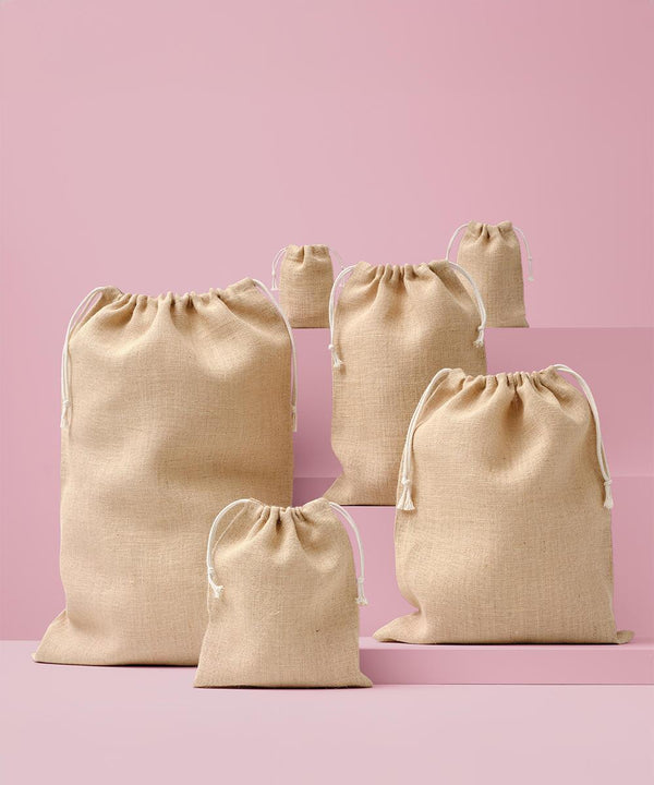 Five Nutshell jute stuff bags in assorted sizes stand upright on pink surfaces with a pink background. Each beige bag features white drawstrings.