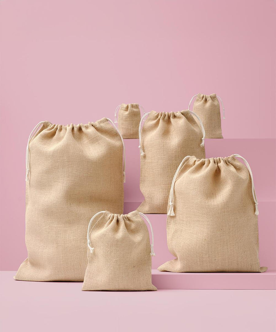 Five Nutshell jute stuff bags in assorted sizes stand upright on pink surfaces with a pink background. Each beige bag features white drawstrings.