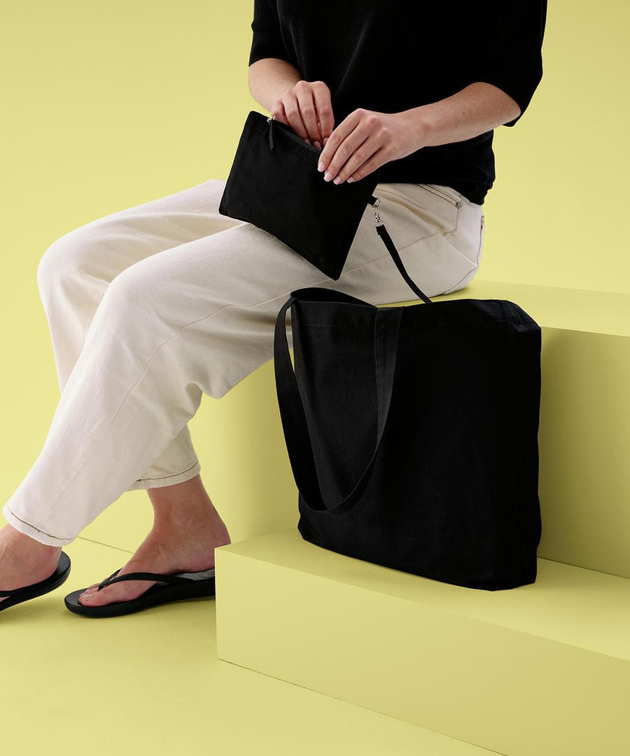 A person sits on pale yellow steps, holding the Nutshell Cruise recycled tote with detachable purse, both in black. They wear a black top, white trousers, and black sandals. The large tote bag is placed beside them; the background and steps are pale yellow.