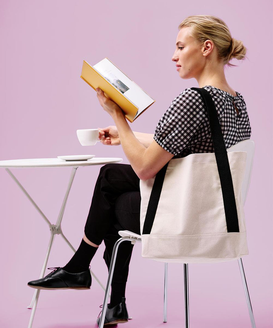A person sits at a white table with a cup and saucer, holding an open book. They wear a black and white chequered top, black trousers, black shoes, and carry the Nutshell Dakota recycled contrast strap tote. The background is pink.
