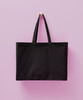 The Nutshell Billie recycled luxe maxi tote, a plain black bag with two handles, hangs on a wooden peg against a light pink background.