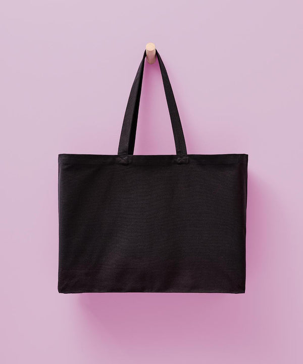 The Nutshell Billie recycled luxe maxi tote, a plain black bag with two handles, hangs on a wooden peg against a light pink background.
