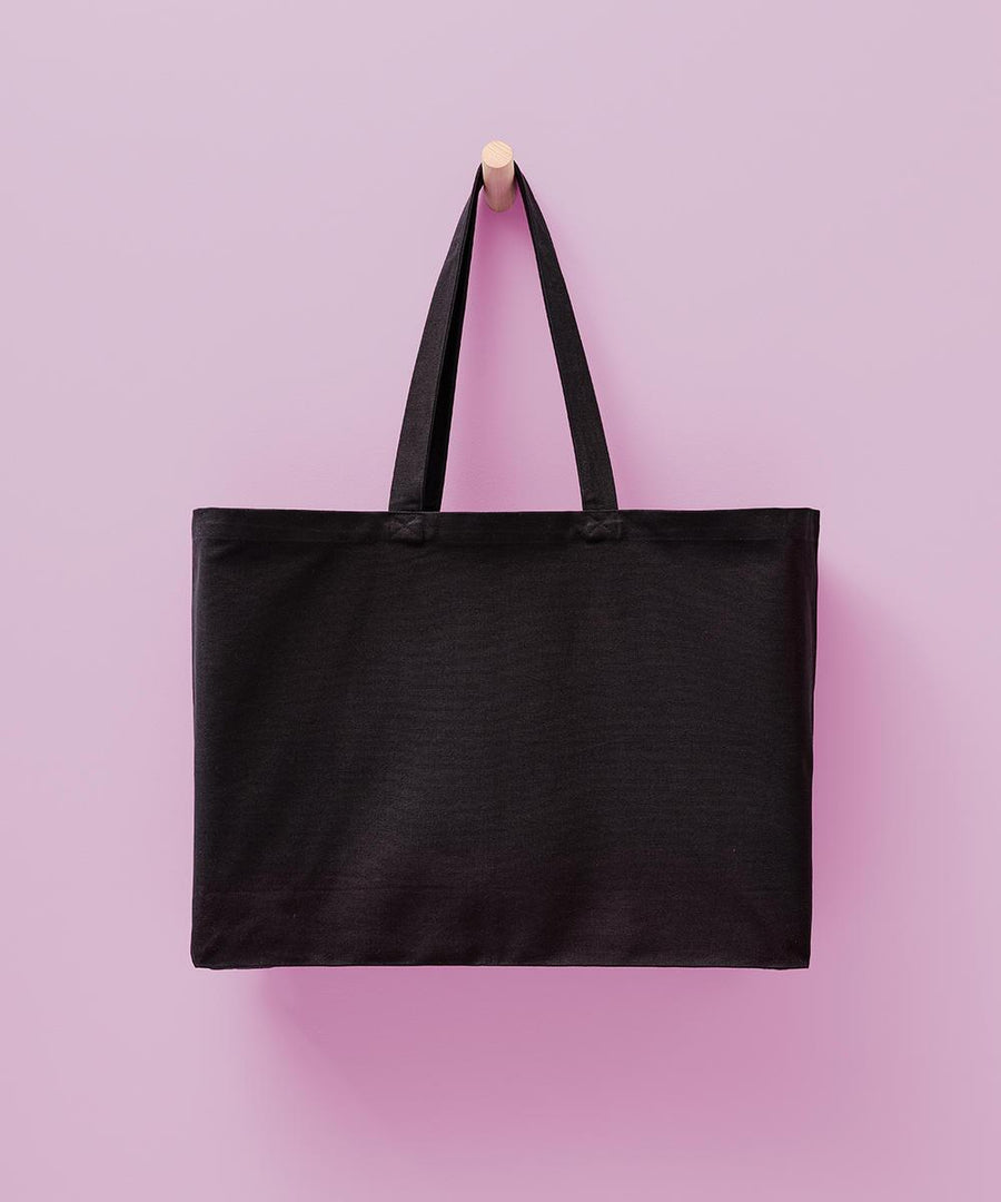 The Nutshell Billie recycled luxe maxi tote, a plain black bag with two handles, hangs on a wooden peg against a light pink background.