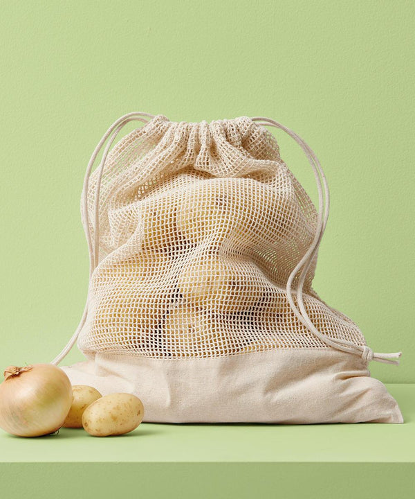A Nutshell cotton mesh bag filled with potatoes sits on a green surface, alongside two potatoes and an onion, all set against a light green background.