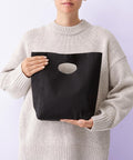 A person in a light oversized knitted jumper holds the Nutshell Frankie recycled premium bag—a black fabric bag with an oval cut-out handle—against a plain light background, their face partially visible.