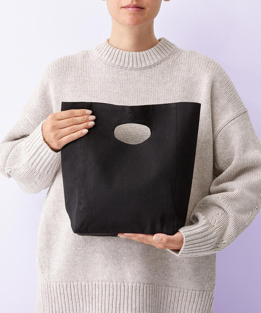 A person in a light oversized knitted jumper holds the Nutshell Frankie recycled premium bag—a black fabric bag with an oval cut-out handle—against a plain light background, their face partially visible.