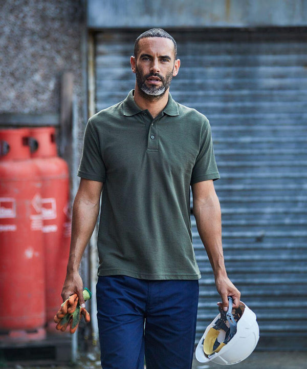 A man in a green ProRTX RX101 and navy pants holds a white helmet and gloves. Two red gas cylinders and a corrugated metal door are in the background.