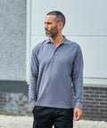 A man with short dark hair and a beard stands outdoors wearing a long-sleeve, grey RX102 polo shirt by ProRTX, looking to his left. The background includes parts of a building and gravel on the ground.