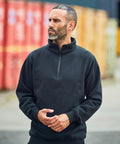 A man with short hair and a beard wears a black RX305 zip-up sweater by ProRTX and stands outdoors near shipping containers, looking to his left.