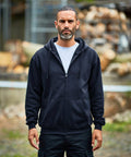 A man with short hair and a beard wearing a black ProRTX RX351 zip-up hoodie and black pants stands outdoors, with a neutral expression on his face. Logs and metal structures are visible in the background.