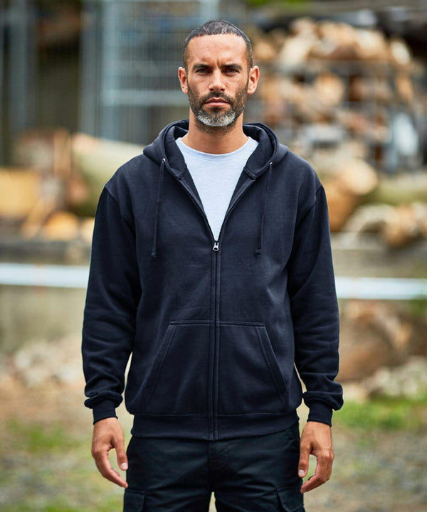 A man with short hair and a beard wearing a black ProRTX RX351 zip-up hoodie and black pants stands outdoors, with a neutral expression on his face. Logs and metal structures are visible in the background.