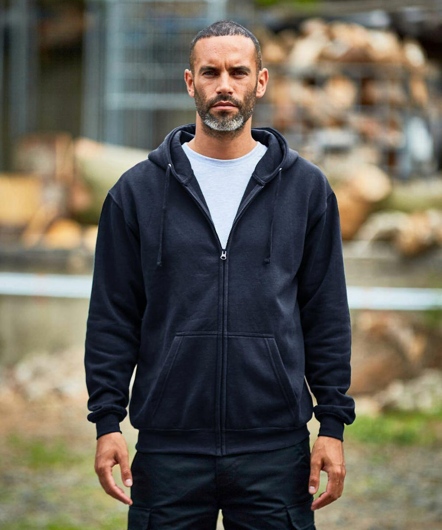 A man with short hair and a beard wearing a black ProRTX RX351 zip-up hoodie and black pants stands outdoors, with a neutral expression on his face. Logs and metal structures are visible in the background.