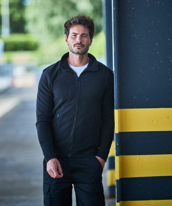 A man wearing a black RX401 by ProRTX jacket and black pants stands against a dark wall with yellow and black stripes. He has one hand in his pocket and is looking slightly towards the camera.