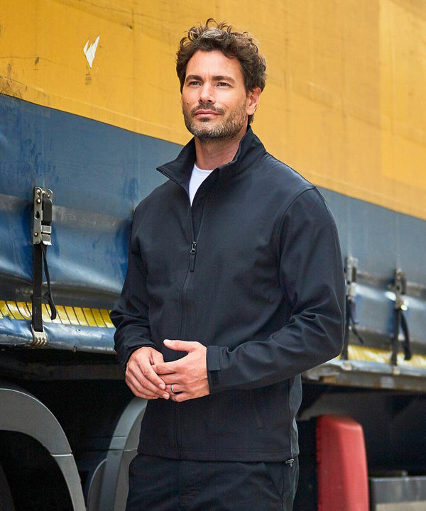 A man with short curly hair and facial hair is wearing a black RX500 jacket by ProRTX and standing outdoors in front of a yellow and blue industrial background.