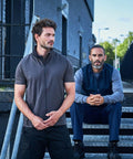 Two men are posing outdoors. One man is standing while the other is sitting on stairs next to a handrail, with a building and sky in the background. Both are wearing casual, short-sleeve tops from the RX550 line by ProRTX.