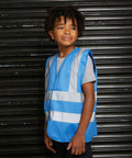 A child wearing a blue and silver reflective ProRTX RX70J safety vest stands in front of a black corrugated metal background.