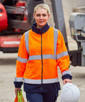 A person wearing a ProRTX RX750 orange high-visibility jacket and holding a white hard hat stands in an industrial area with machinery in the background.