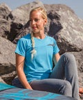 A person with a braid in a blue Spiro S182F shirt and gray leggings sits on a rock outdoors.