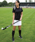 A woman in a black and white sports outfit holds a Spiro S261F field hockey stick on a grassy field, with a stadium in the background.