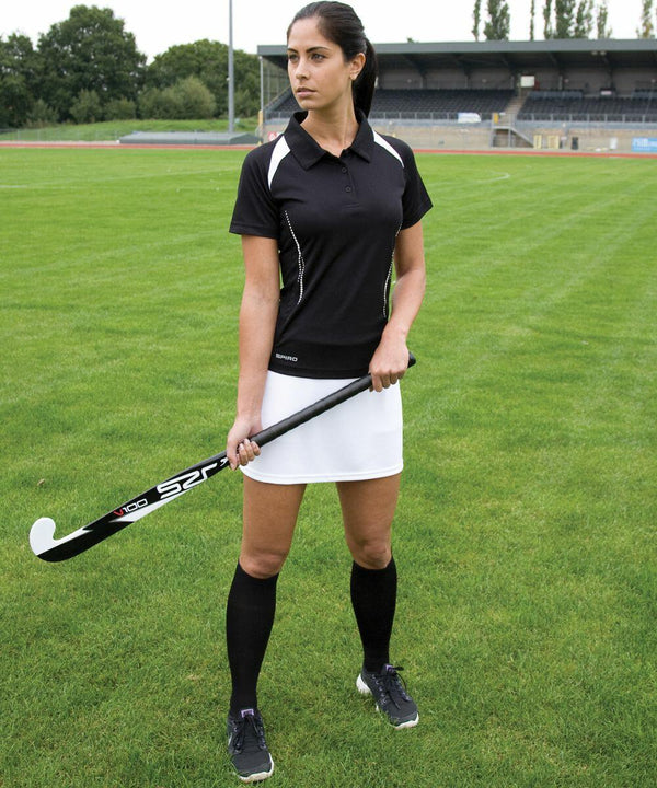 A woman in a black and white sports outfit holds a Spiro S261F field hockey stick on a grassy field, with a stadium in the background.