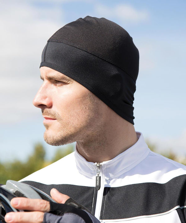 A man outdoors wearing a Spiro bikewear winter hat and a white jacket with black stripes looks to the side, holding an object in his gloved hand. The bright background is slightly blurred.