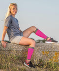 A person with long blonde hair sits on a stone ledge outdoors, wearing a gray shirt, beige shorts, pink knee-high socks, and black Spiro S290X athletic shoes with pink accents.