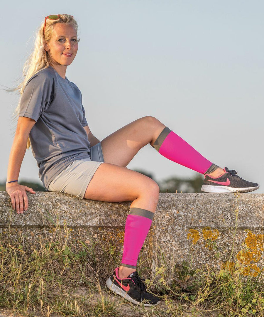 A person with long blonde hair sits on a stone ledge outdoors, wearing a gray shirt, beige shorts, pink knee-high socks, and black Spiro S290X athletic shoes with pink accents.