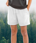 A person stands with hands in the pockets of Spiro Recycled junior shorts and a dark shirt, against a backdrop of a misty forest with green trees.
