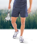 A person wearing Spiro Recycled men’s shorts, a matching shirt, and white trainers stands with a lacrosse stick. Behind them, a misty forest rises above the trees.