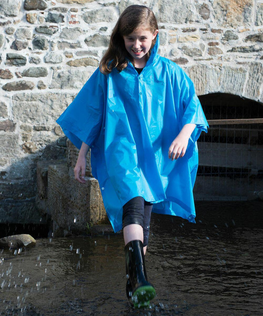 A person wearing a blue Splashmac SC019 poncho and black boots walks through a shallow water stream in front of a stone wall.