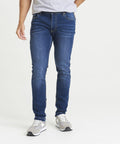 A person wearing a gray t-shirt, blue jeans (SD004 by AWDis So Denim), and gray sneakers is standing against a plain white background. Their hands are partially in their pockets.