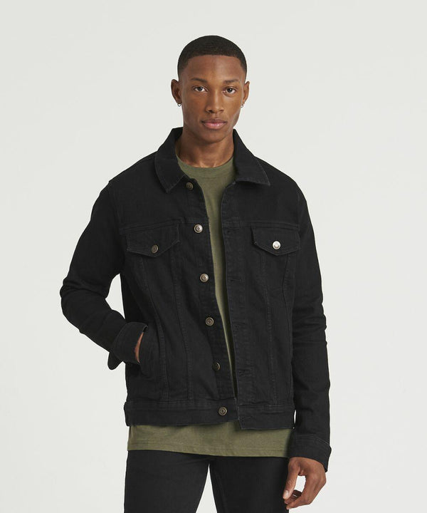 A person stands facing the camera, wearing a black AWDis So Denim SD060 denim jacket over a green shirt, with one hand in their pocket.